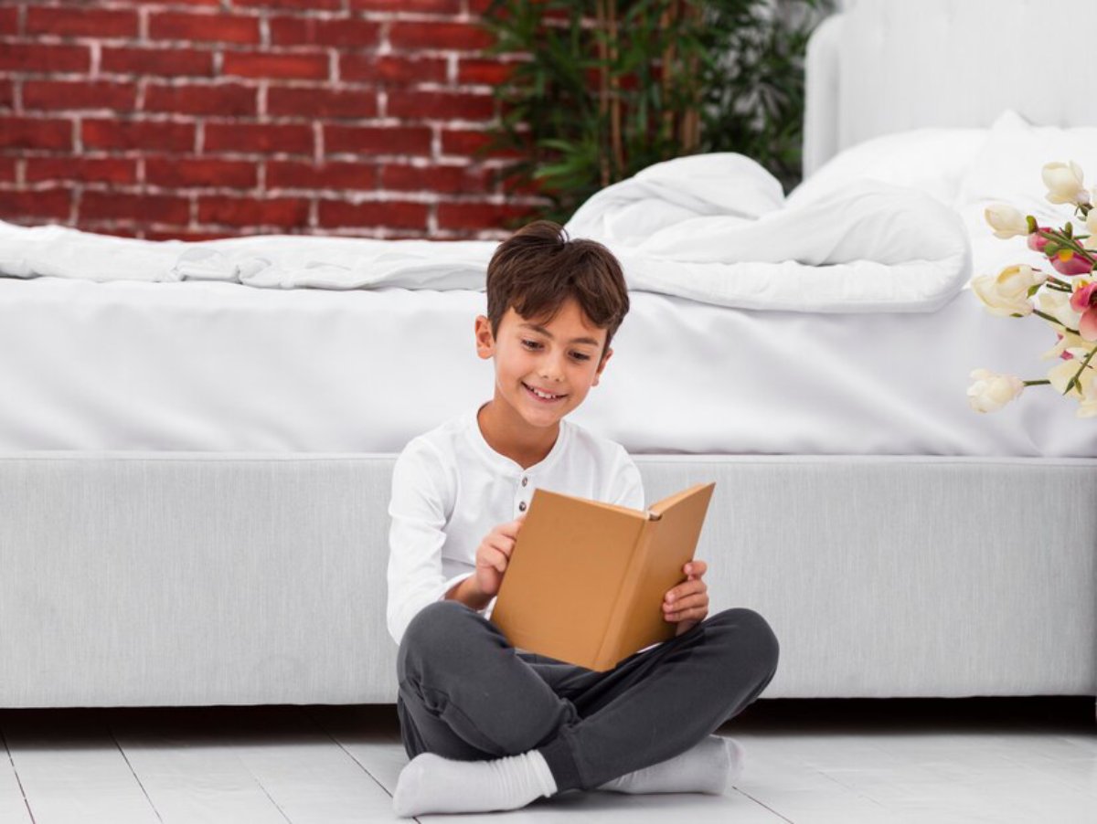 A child sits cross-legged on the floor, reading a book in a bright room with a brick wall and a neatly made bed in the background.