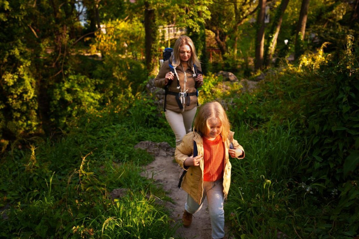 A woman and a child hike along a sunlit path surrounded by lush greenery. The scene captures a peaceful outdoor adventure.