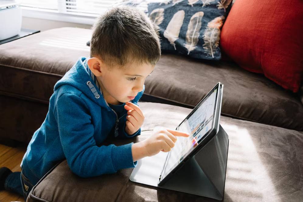 A young child in a blue hoodie interacts with a tablet on a brown couch, surrounded by colorful cushions and natural light.