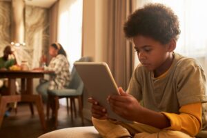 Young boy focused on using a tablet, with family members dining in the background at home.