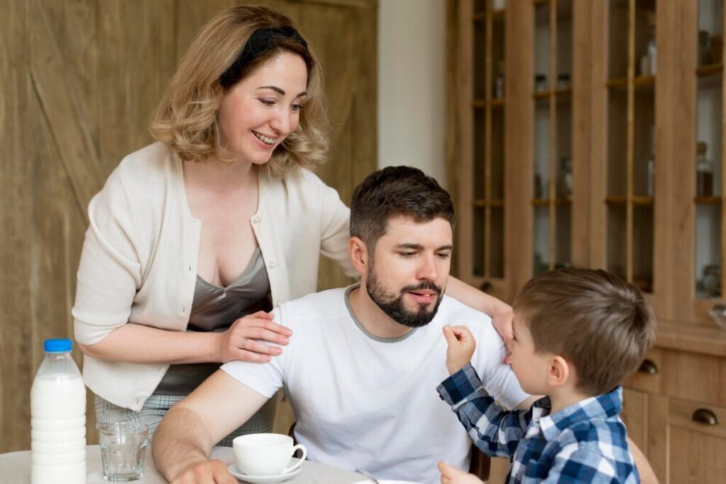 A family of three sits around a table with a cup and glass, sharing a moment in a cozy, well-decorated kitchen.