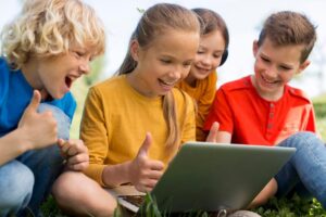 Four children sit on the grass, smiling and giving thumbs up while excitedly looking at a laptop in a sunny outdoor setting.