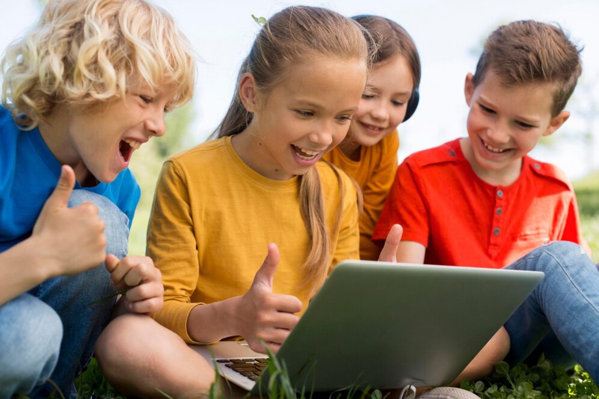 Four children sit on the grass, smiling and giving thumbs up while excitedly looking at a laptop in a sunny outdoor setting.