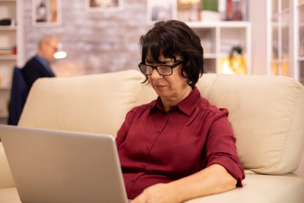 A woman in a burgundy blouse sits on a beige couch, focused on her laptop, with a softly lit background of shelves and decor.