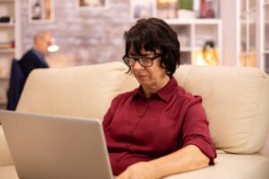 A woman in a burgundy blouse sits on a beige couch, focused on her laptop, with a softly lit background of shelves and decor.