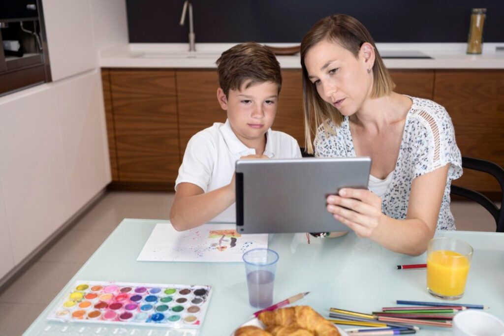 A woman and a boy engage with a tablet at a kitchen table, surrounded by art supplies and snacks. A colorful painting is displayed nearby.
