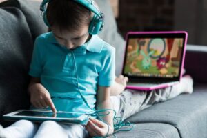 A child in a turquoise shirt sits on a couch, using a tablet while watching cartoons on a pink laptop. Headphones cover their ears.