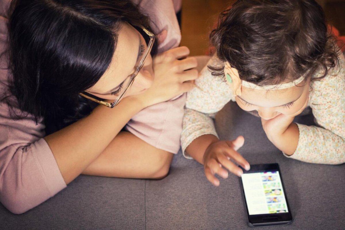 A woman and a child sit together, focused on a smartphone displaying colorful content while leaning on a gray surface.