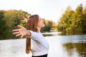 Young woman joyfully stretching her arms by a scenic river surrounded by trees.
