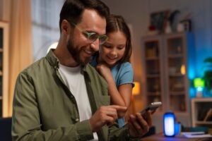 A person in a green shirt interacts with a smartphone while a child leans on them, surrounded by a cozy, softly lit room.