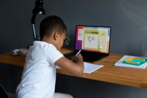A child in a white shirt writes in a notebook while engaged with a math lesson on a laptop, surrounded by colorful stationery.