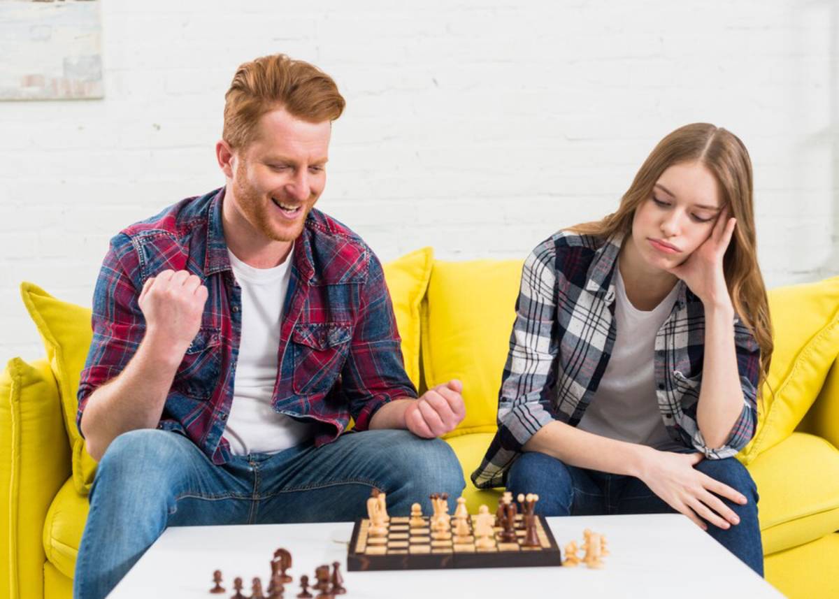 A man enthusiastically plays chess with a woman who appears bored, both sitting on a yellow couch.